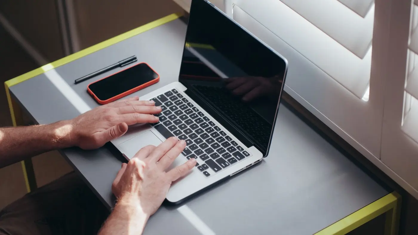 Mani che lavorano al portatile su una scrivania vicino a una finestra, con smartphone e penna accanto.