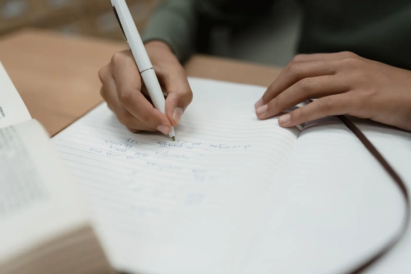 Primo piano di una mano che scrive con una penna bianca su un quaderno a righe aperto.