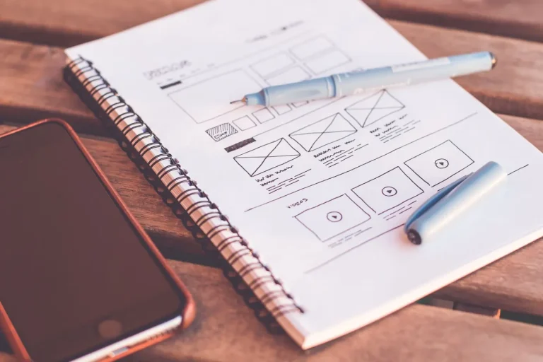 Quaderno con schizzi di wireframe di un sito web accanto a uno smartphone spento su un tavolo di legno.