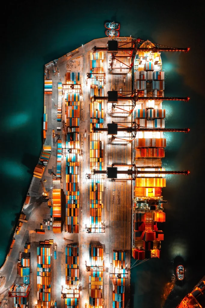 Veduta aerea notturna di un terminal portuale con gru e nave carica di container colorati illuminati.