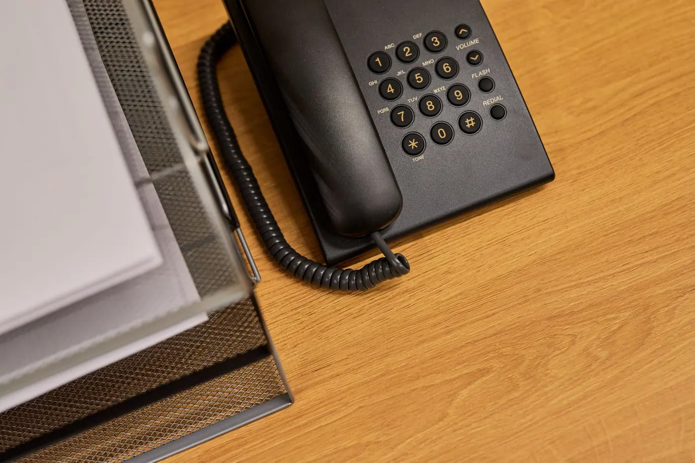 Telefono da scrivania nero con cornetta e filo a spirale su una scrivania in legno, vicino a un portadocumenti in rete metallica.