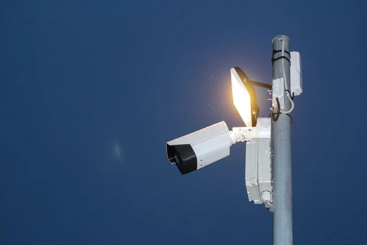 Telecamera di sicurezza bullet bianca e nera montata su un palo accanto a un faro illuminato, contro un cielo blu notte.