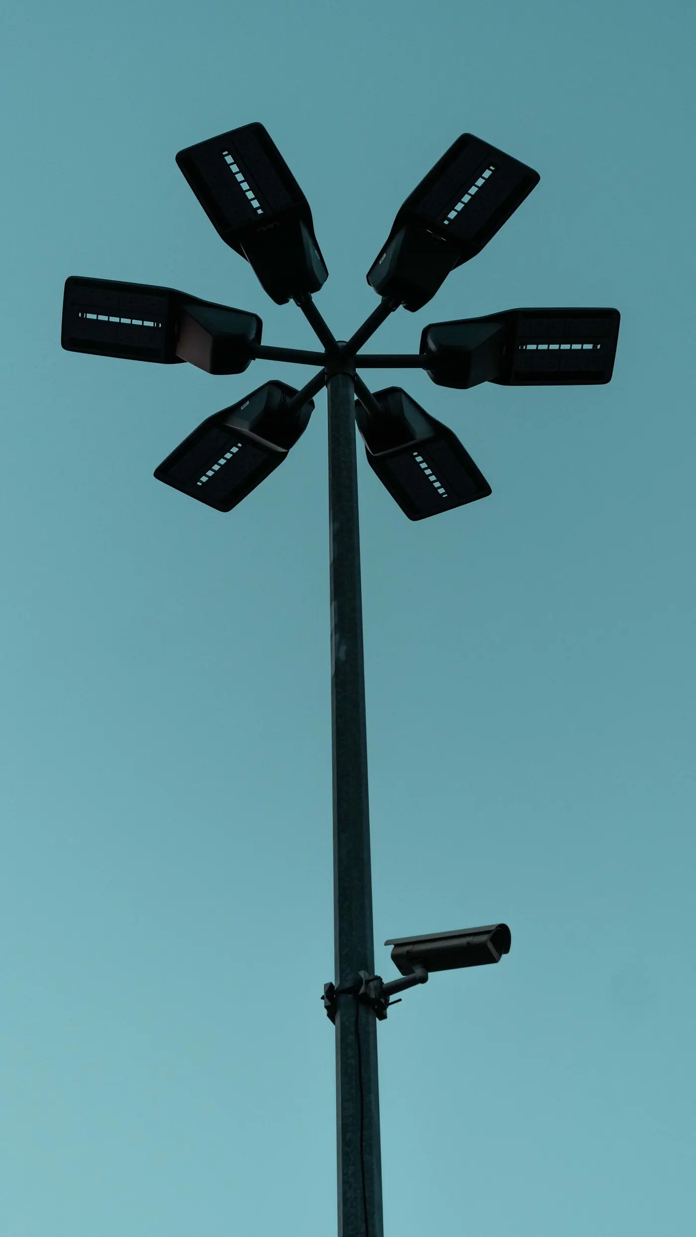 Telecamera di sorveglianza fissata a un alto palo con proiettori a LED, in silhouette contro un cielo azzurro-verde del crepuscolo.